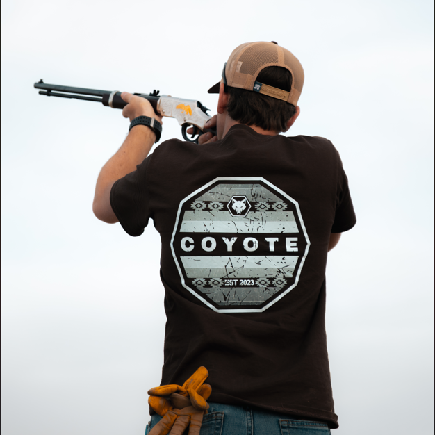 Person wearing a 'Coyote' t-shirt holding a rifle against a clear sky.
